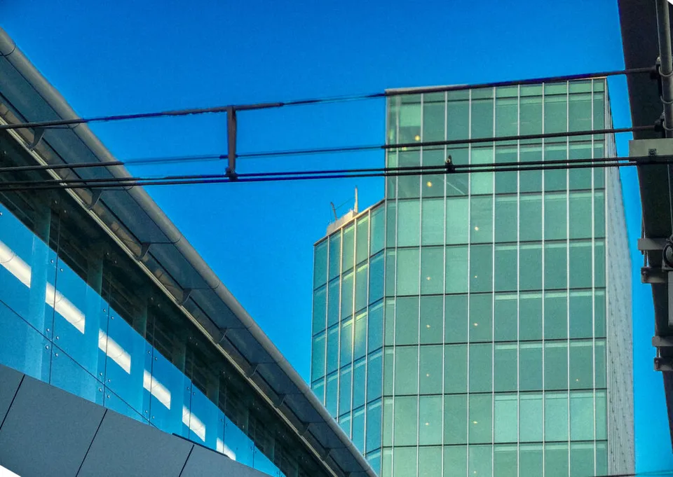 An abstract, geometric composition focusing on modern urban architecture. A glass-walled pedestrian bridge or elevated walkway is visible on the left, angled towards the upper right, reflecting light. Behind it and to the right is a tall, square, glass skyscraper with a pale green tint against a bright blue sky. Overhead wires cross the top of the frame.