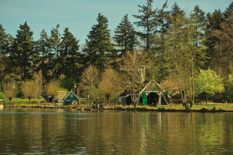 A wide landscape shot shows a serene lake with traditional green wooden buildings and a small windmill along the grassy shore. Tall evergreen trees stand behind the buildings, and their reflections shimmer in the calm, yellowish-toned water in the foreground.
