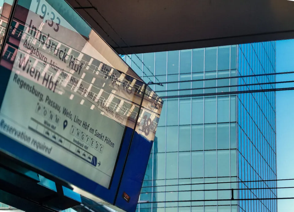 A close-up, angled view of an outdoor train station departure board screen listing destinations like Innsbruck Hbf, Wien Hbf, and Augsburg. The bright screen reflects a nearby modern glass skyscraper, which fills the background of the image with a blue tint.