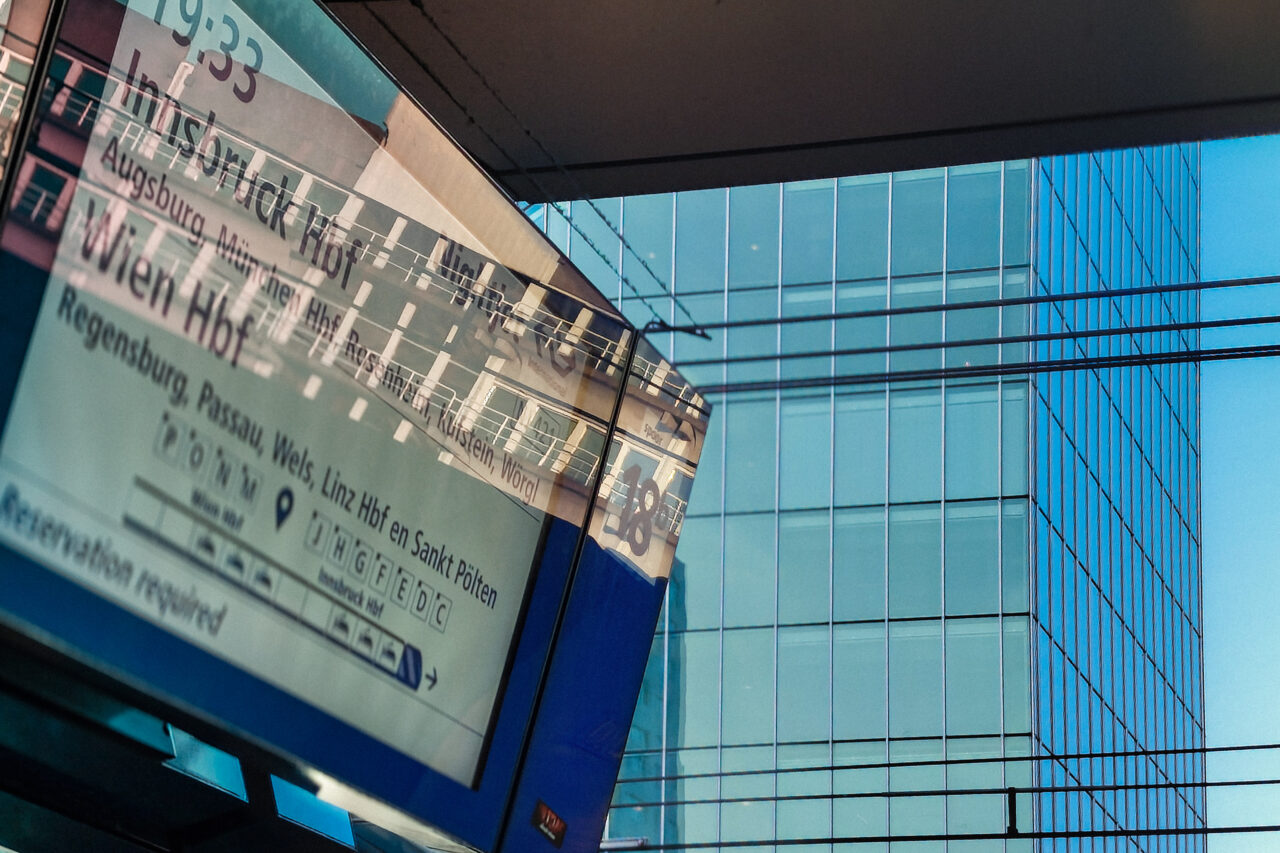 A close-up, angled view of an outdoor train station departure board screen listing destinations like Innsbruck Hbf, Wien Hbf, and Augsburg. The bright screen reflects a nearby modern glass skyscraper, which fills the background of the image with a blue tint.