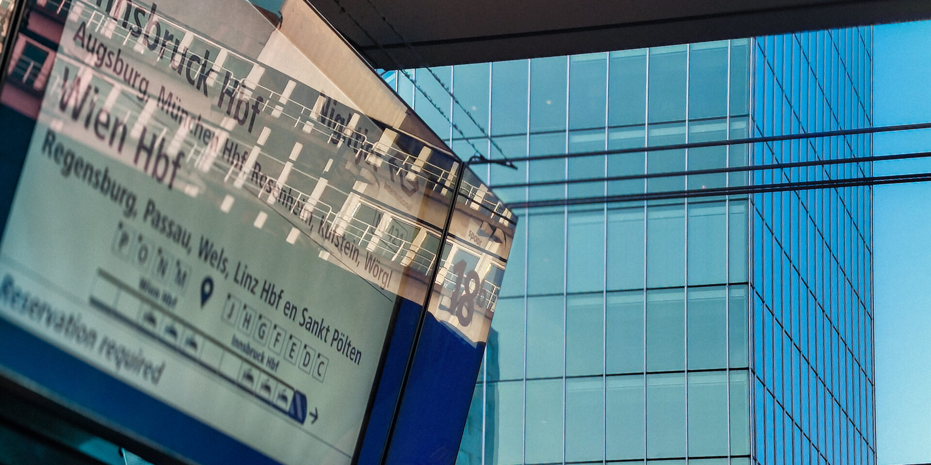 A close-up, angled view of an outdoor train station departure board screen listing destinations like Innsbruck Hbf, Wien Hbf, and Augsburg. The bright screen reflects a nearby modern glass skyscraper, which fills the background of the image with a blue tint.