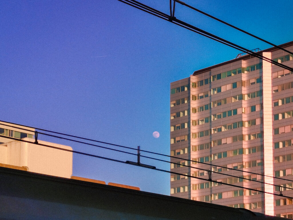 A sunset or twilight urban landscape. The sky is a gradient of deep blue and purple. A tall, rectangular apartment or office building on the right is bathed in warm, orange light from the setting sun. A small, white half-moon is visible in the middle sky, positioned between the building and the silhouette of overhead railway wires and a lower concrete structure.