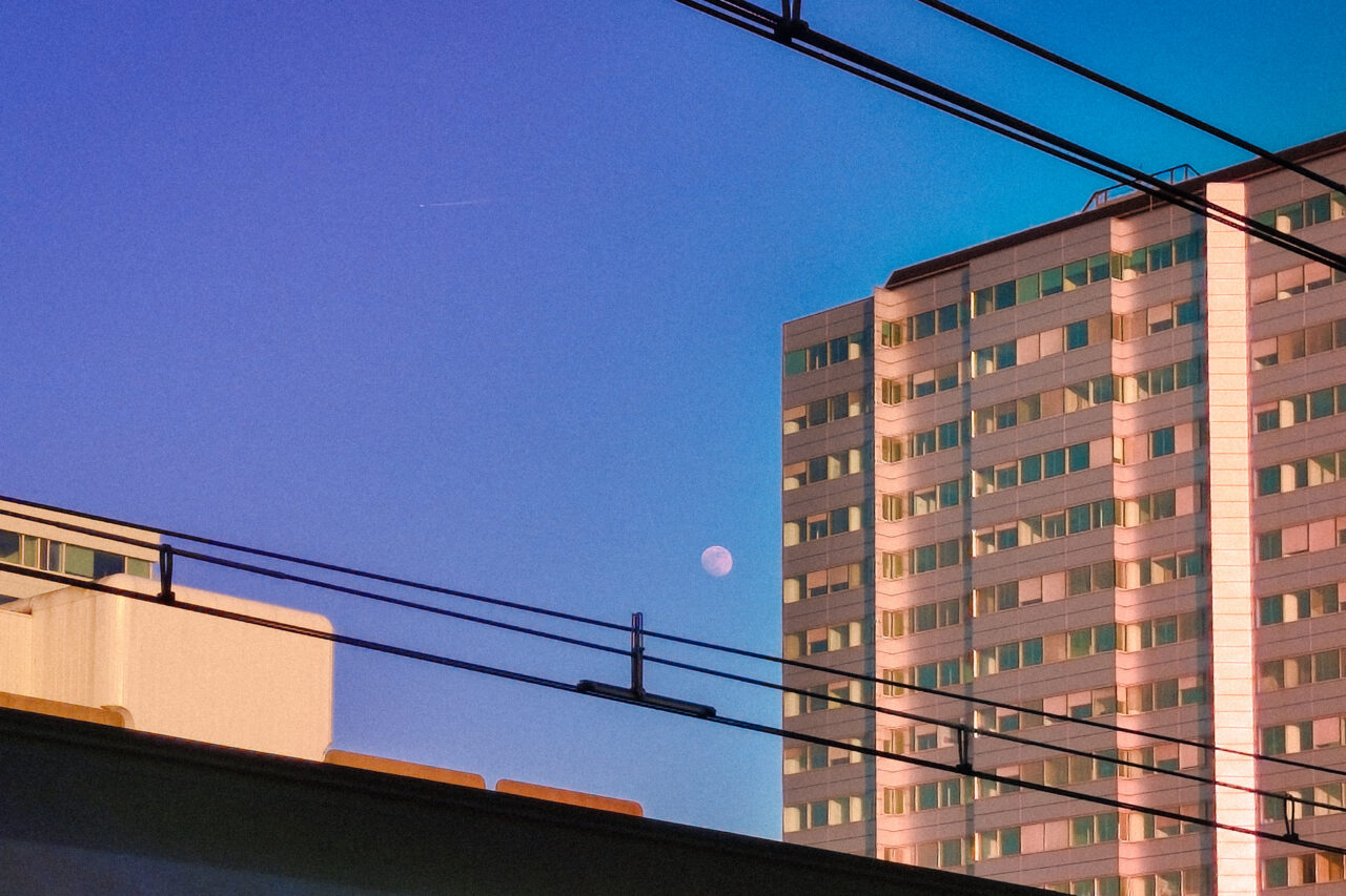A sunset or twilight urban landscape. The sky is a gradient of deep blue and purple. A tall, rectangular apartment or office building on the right is bathed in warm, orange light from the setting sun. A small, white half-moon is visible in the middle sky, positioned between the building and the silhouette of overhead railway wires and a lower concrete structure.
