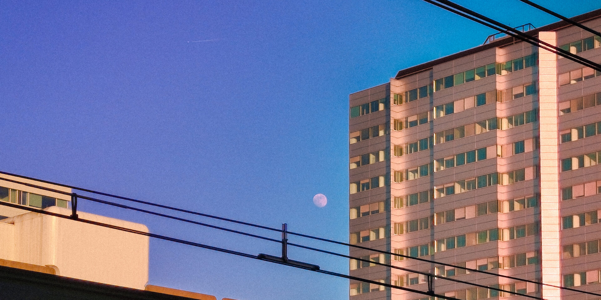 A sunset or twilight urban landscape. The sky is a gradient of deep blue and purple. A tall, rectangular apartment or office building on the right is bathed in warm, orange light from the setting sun. A small, white half-moon is visible in the middle sky, positioned between the building and the silhouette of overhead railway wires and a lower concrete structure.