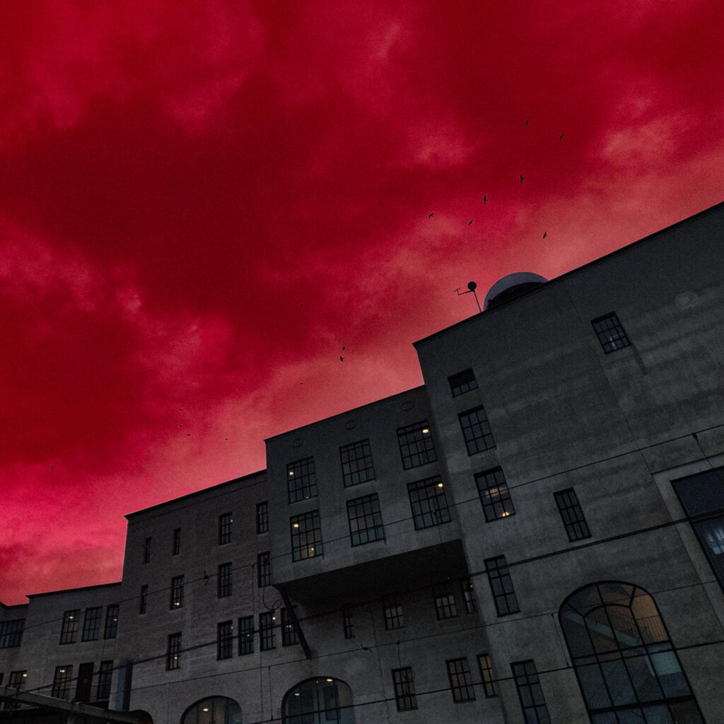 A vertical, low-angle shot of a large, imposing, gray concrete or stone building, likely an old factory or industrial warehouse, against a dramatically vivid, artificially colored deep red sky. The building features multiple rectangular and arched windows and is viewed from below, emphasizing its height and shadow. Several small black bird silhouettes are visible against the fiery red clouds.