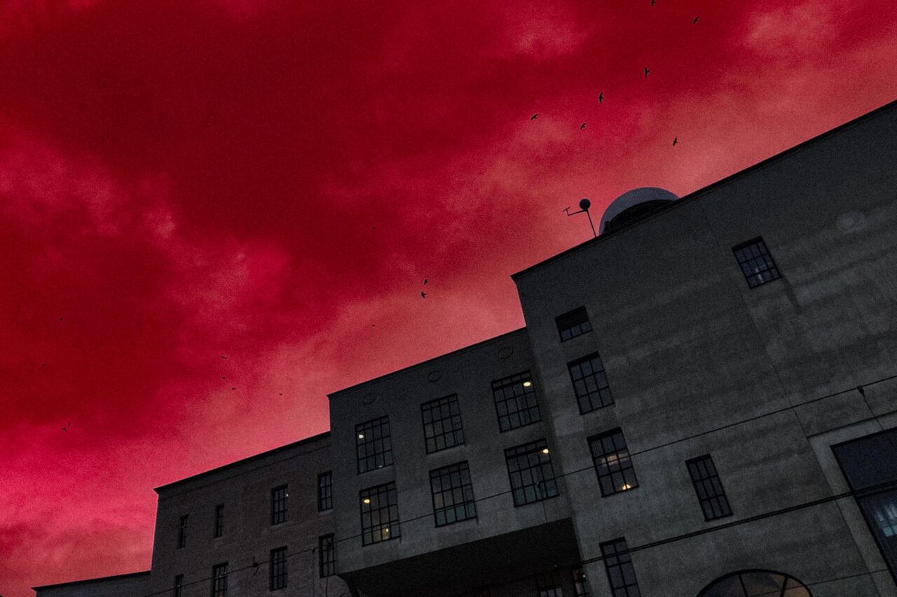 A vertical, low-angle shot of a large, imposing, gray concrete or stone building, likely an old factory or industrial warehouse, against a dramatically vivid, artificially colored deep red sky. The building features multiple rectangular and arched windows and is viewed from below, emphasizing its height and shadow. Several small black bird silhouettes are visible against the fiery red clouds.