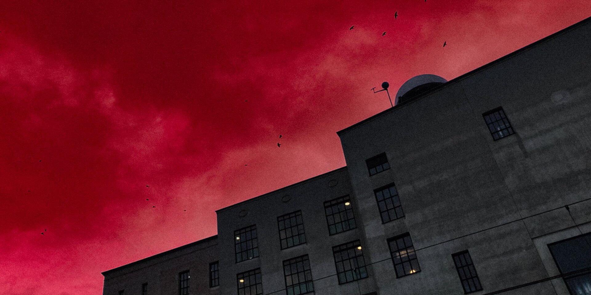 A vertical, low-angle shot of a large, imposing, gray concrete or stone building, likely an old factory or industrial warehouse, against a dramatically vivid, artificially colored deep red sky. The building features multiple rectangular and arched windows and is viewed from below, emphasizing its height and shadow. Several small black bird silhouettes are visible against the fiery red clouds.