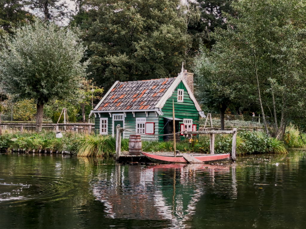 A small, traditional green wooden cottage with a red-tiled roof sits at the edge of a pond or canal. The house has white window frames and a chimney. A small, red wooden boat is tied to a wooden dock directly in front of the house, next to a wooden barrel. The water in the foreground reflects the house and surrounding greenery, and the background is densely packed with various trees under an overcast sky.