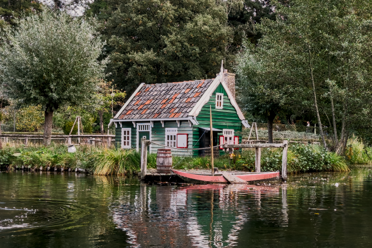A small, traditional green wooden cottage with a red-tiled roof sits at the edge of a pond or canal. The house has white window frames and a chimney. A small, red wooden boat is tied to a wooden dock directly in front of the house, next to a wooden barrel. The water in the foreground reflects the house and surrounding greenery, and the background is densely packed with various trees under an overcast sky.