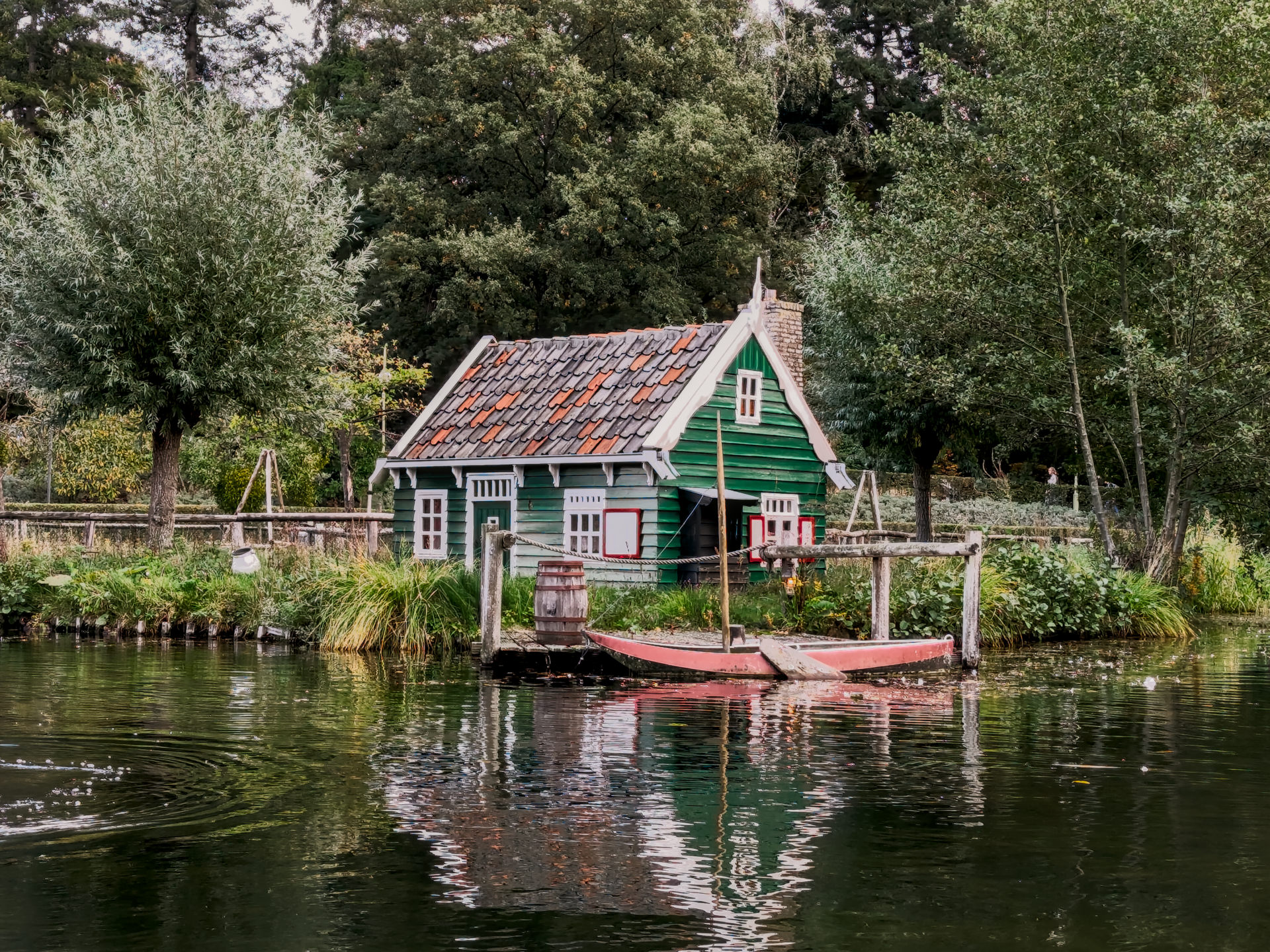 A small, traditional green wooden cottage with a red-tiled roof sits at the edge of a pond or canal. The house has white window frames and a chimney. A small, red wooden boat is tied to a wooden dock directly in front of the house, next to a wooden barrel. The water in the foreground reflects the house and surrounding greenery, and the background is densely packed with various trees under an overcast sky.