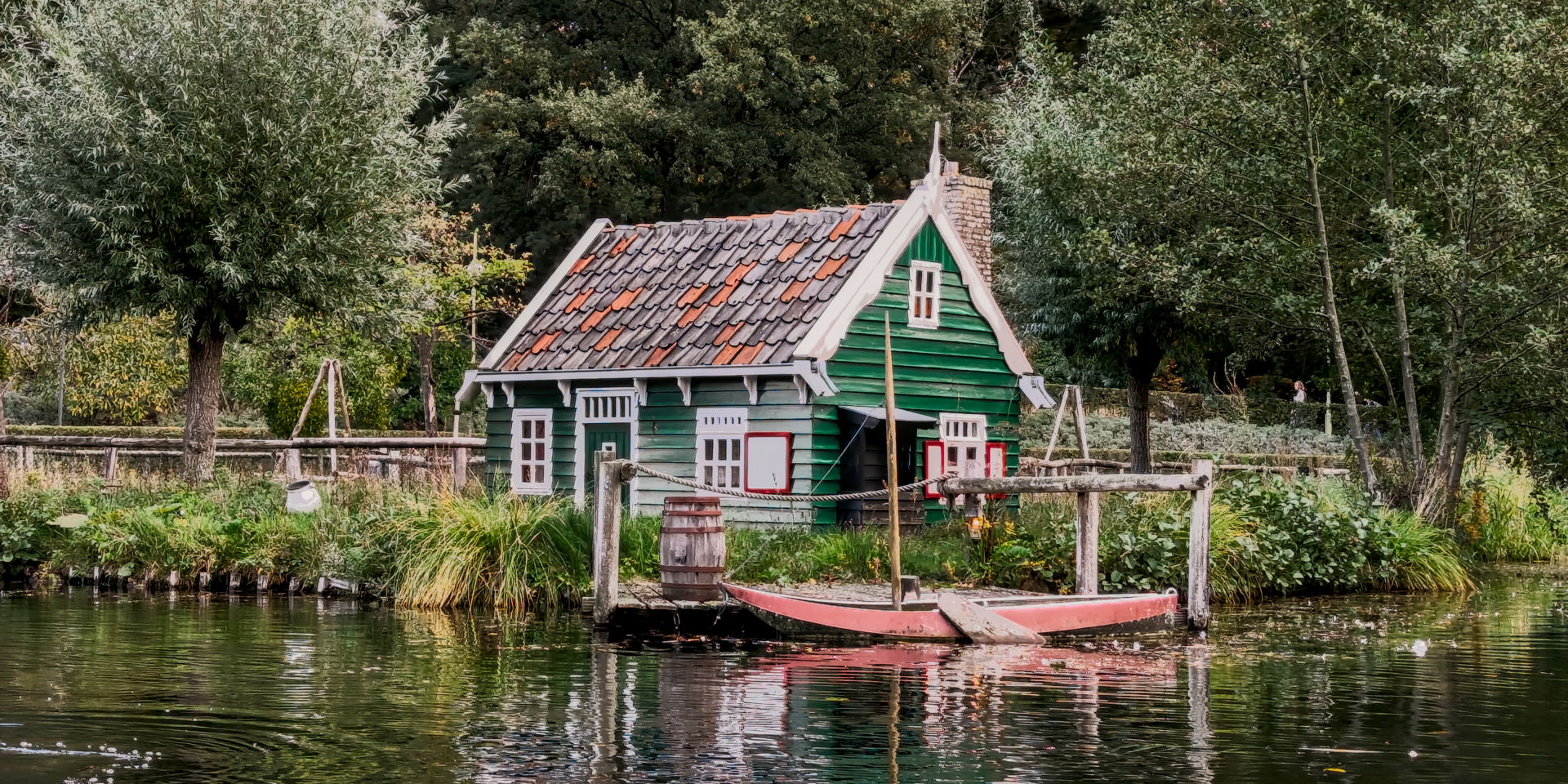A small, traditional green wooden cottage with a red-tiled roof sits at the edge of a pond or canal. The house has white window frames and a chimney. A small, red wooden boat is tied to a wooden dock directly in front of the house, next to a wooden barrel. The water in the foreground reflects the house and surrounding greenery, and the background is densely packed with various trees under an overcast sky.