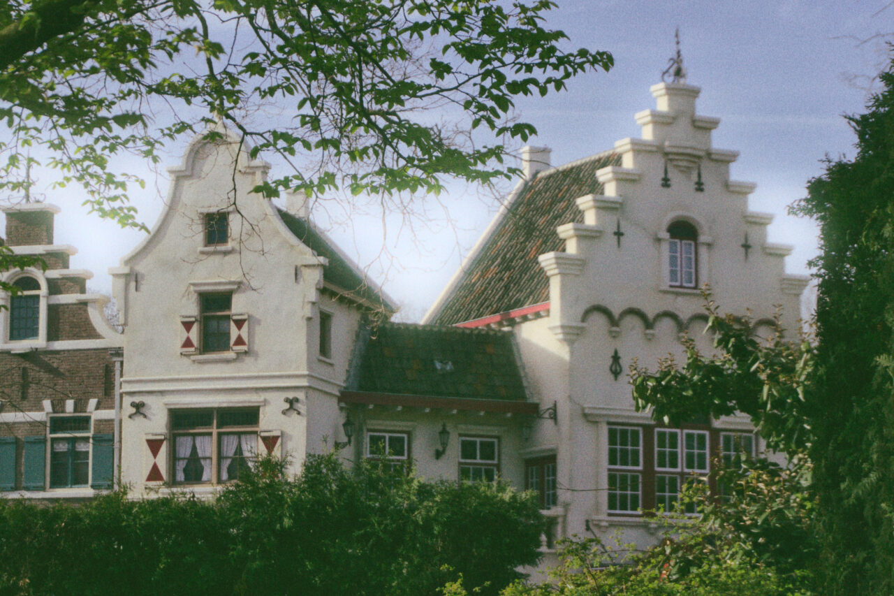 A vertical shot of white, traditional Dutch-style buildings with ornate stepped gables under a soft blue sky. The houses feature dark-framed windows with small red and white shutters and are partially obscured by lush green trees in the foreground. Branches with bright green leaves frame the top left corner of the image, adding a natural border to the architectural scene. The photo has a slightly grainy, film-like texture with soft sunlight hitting the white facades of the buildings.