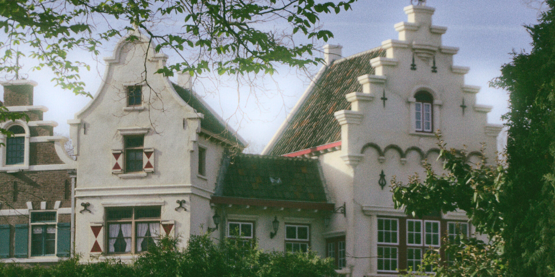 A vertical shot of white, traditional Dutch-style buildings with ornate stepped gables under a soft blue sky. The houses feature dark-framed windows with small red and white shutters and are partially obscured by lush green trees in the foreground. Branches with bright green leaves frame the top left corner of the image, adding a natural border to the architectural scene. The photo has a slightly grainy, film-like texture with soft sunlight hitting the white facades of the buildings.