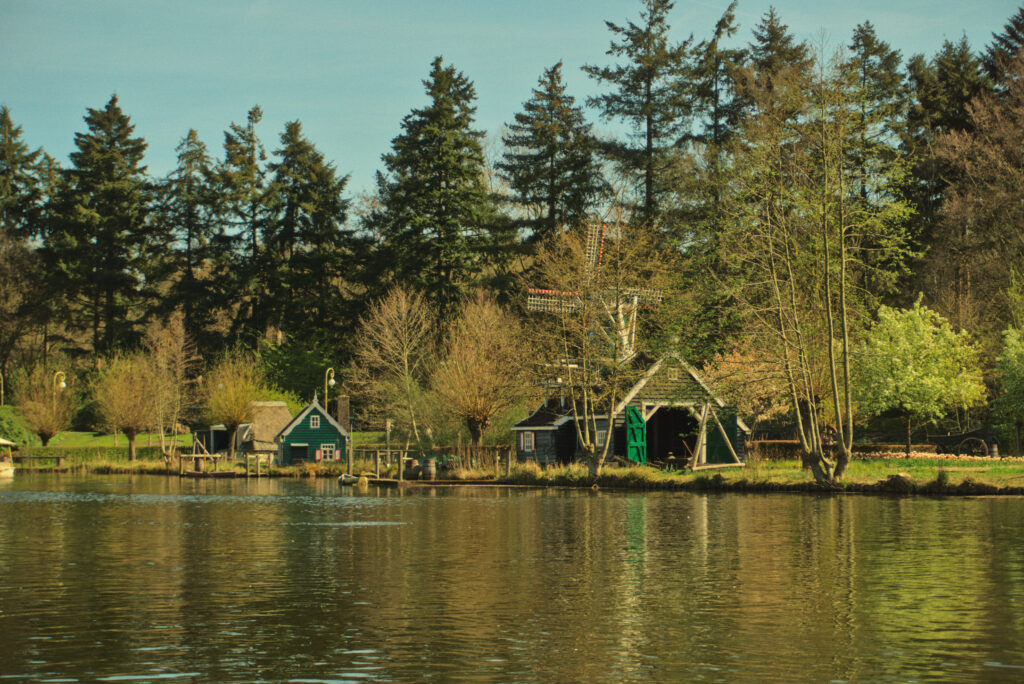 A wide landscape shot shows a serene lake with traditional green wooden buildings and a small windmill along the grassy shore. Tall evergreen trees stand behind the buildings, and their reflections shimmer in the calm, yellowish-toned water in the foreground.