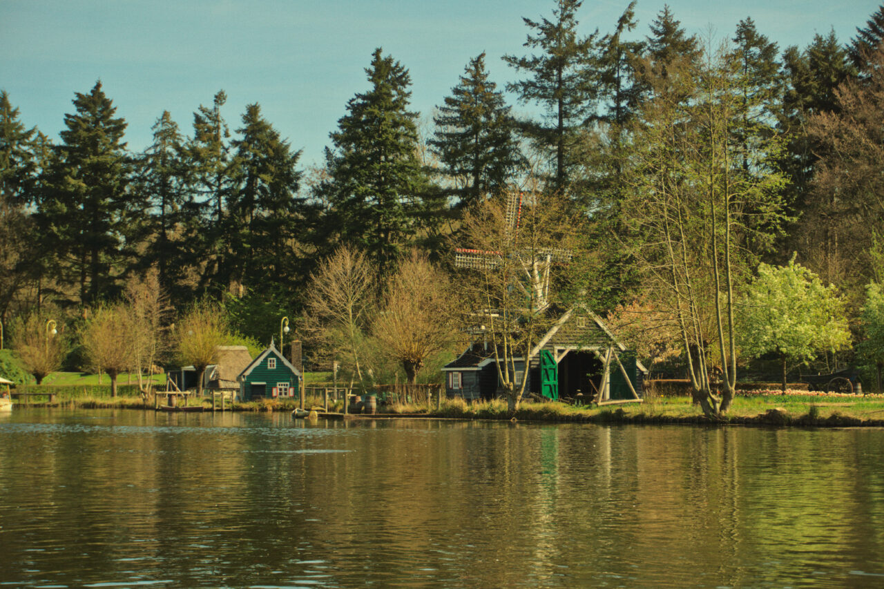 A wide landscape shot shows a serene lake with traditional green wooden buildings and a small windmill along the grassy shore. Tall evergreen trees stand behind the buildings, and their reflections shimmer in the calm, yellowish-toned water in the foreground.