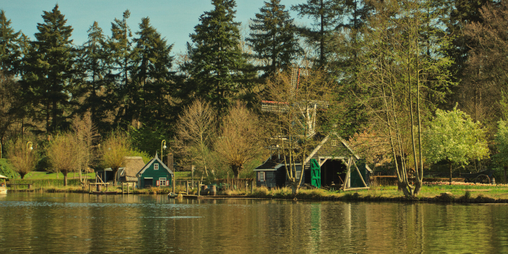 A wide landscape shot shows a serene lake with traditional green wooden buildings and a small windmill along the grassy shore. Tall evergreen trees stand behind the buildings, and their reflections shimmer in the calm, yellowish-toned water in the foreground.