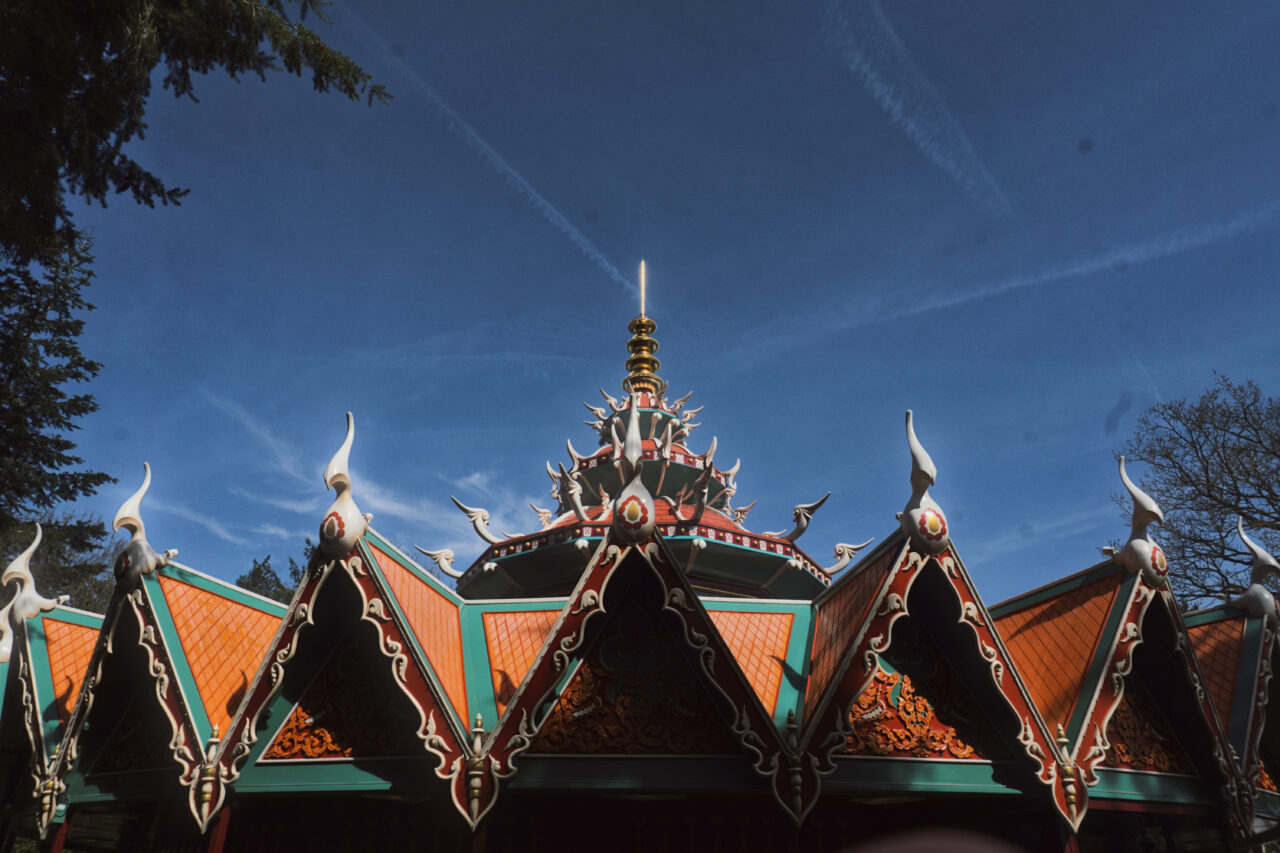 A low-angle, close-up shot focuses on the intricate, multi-tiered roof of a Thai-style structure with orange tiles and green trim. The roof is decorated with white ornamental spikes and gold carvings, set against a deep blue sky with thin white clouds.