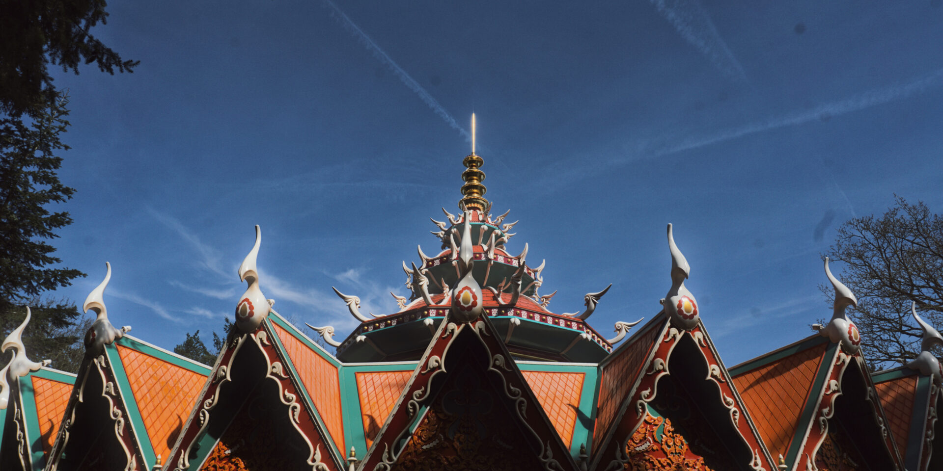 A low-angle, close-up shot focuses on the intricate, multi-tiered roof of a Thai-style structure with orange tiles and green trim. The roof is decorated with white ornamental spikes and gold carvings, set against a deep blue sky with thin white clouds.